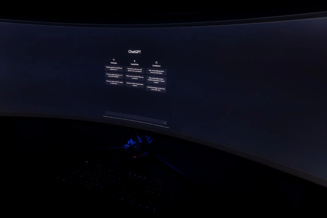 A curved monitor in a dark room showing an AI chat interface, the free tool that competes with any business model built on packaged information