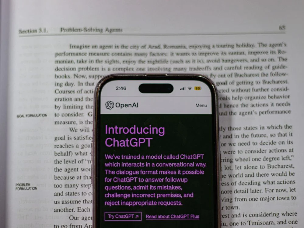 Phone showing an AI product announcement resting on a textbook page about problem-solving agents, the gap between AI tools and a cloned expert methodology