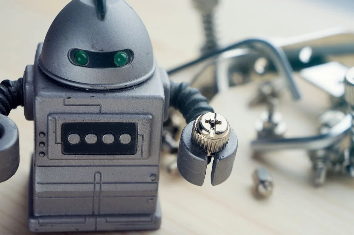 Small metal robot holding a screw on a workbench, illustrating the part of an expert methodology a machine can carry and the part it cannot