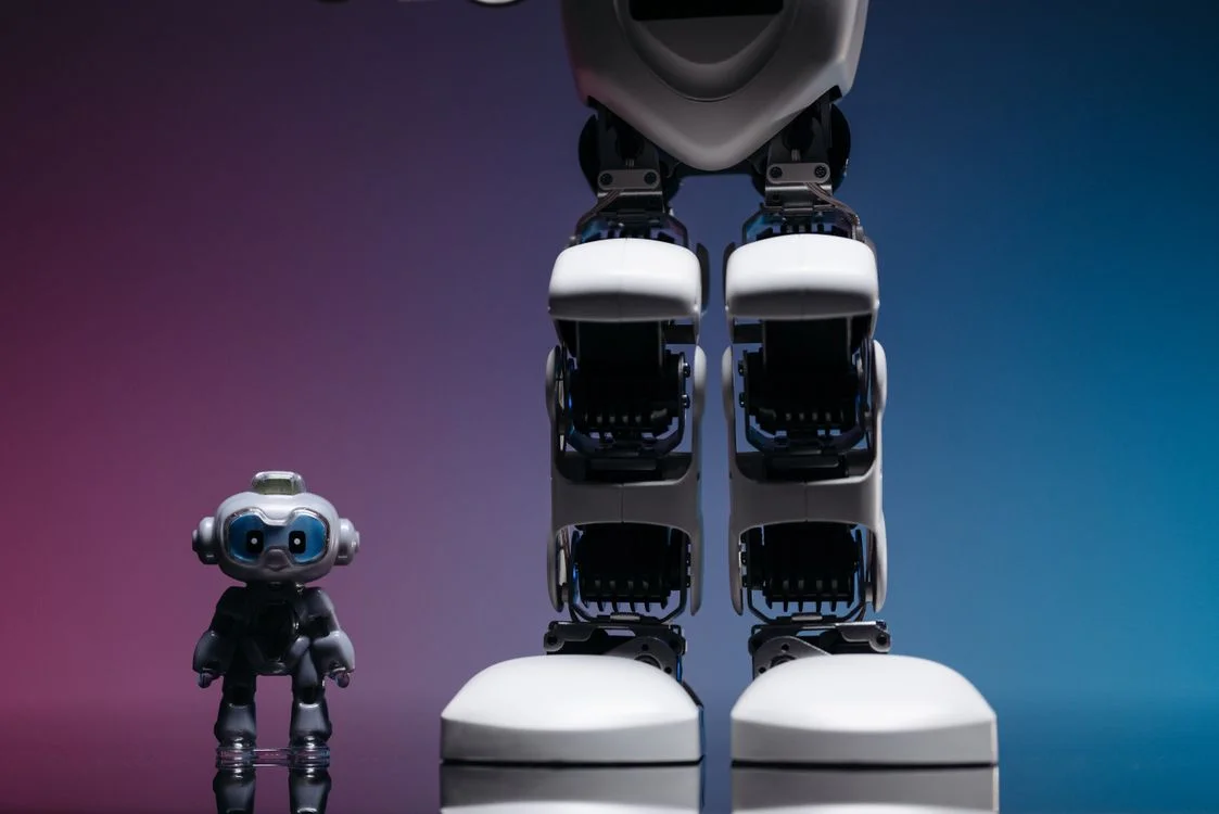 A large humanoid robot standing beside a small toy robot, representing the gap between tool-level AI and system-level AI for agency operations