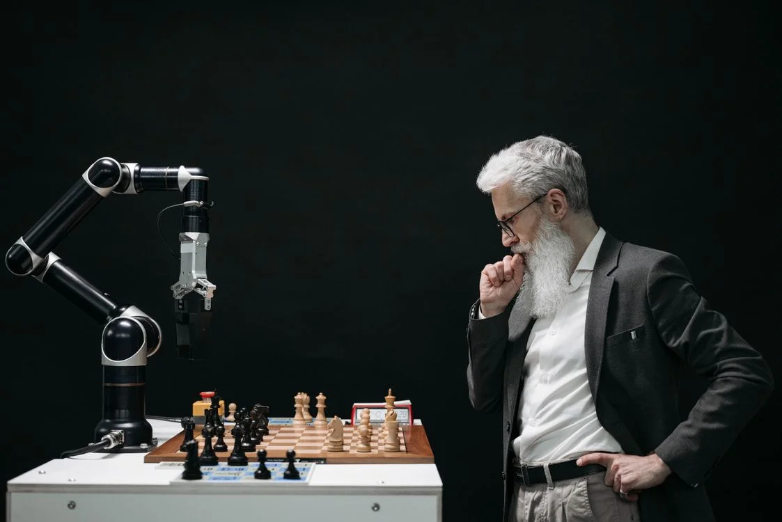 A consultant in a suit studying a chessboard against a robotic arm, representing the judgment work a consultant keeps while an AI agent handles the routine