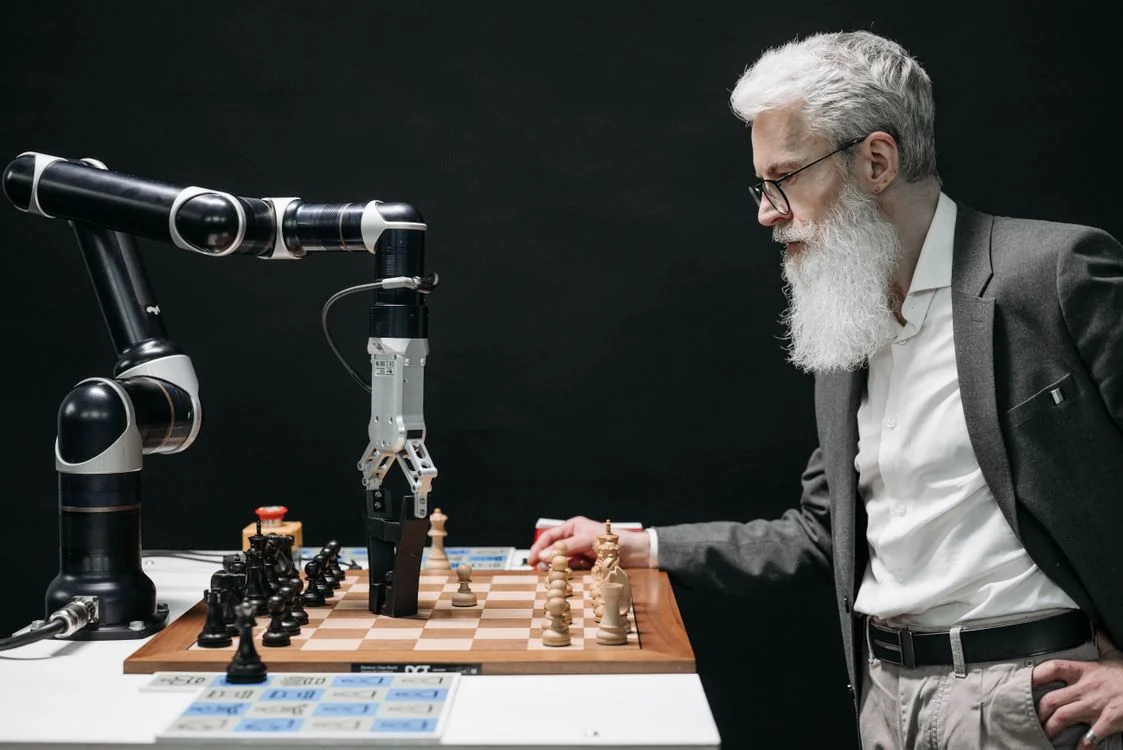 A consultant reaching to move a chess piece against a robotic arm, representing a consultant directing the output while the AI agent executes
