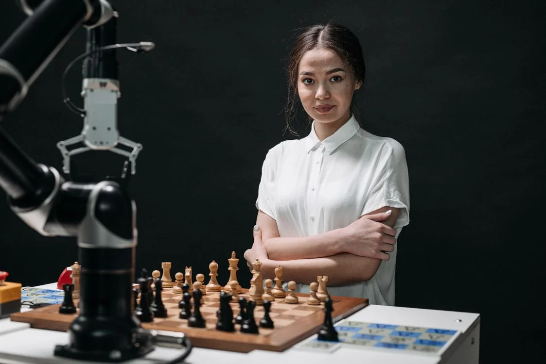 A woman with arms crossed standing beside a chessboard and a black robotic arm, representing the human judgment of deciding when Microsoft Copilot is safe for client work