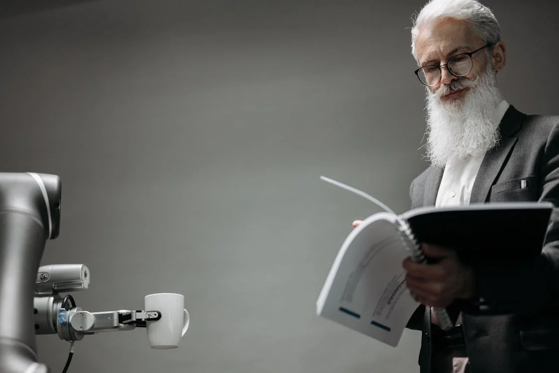 Robotic arm extending coffee to a man reviewing documents — AI assisting the human expert, not replacing their judgment